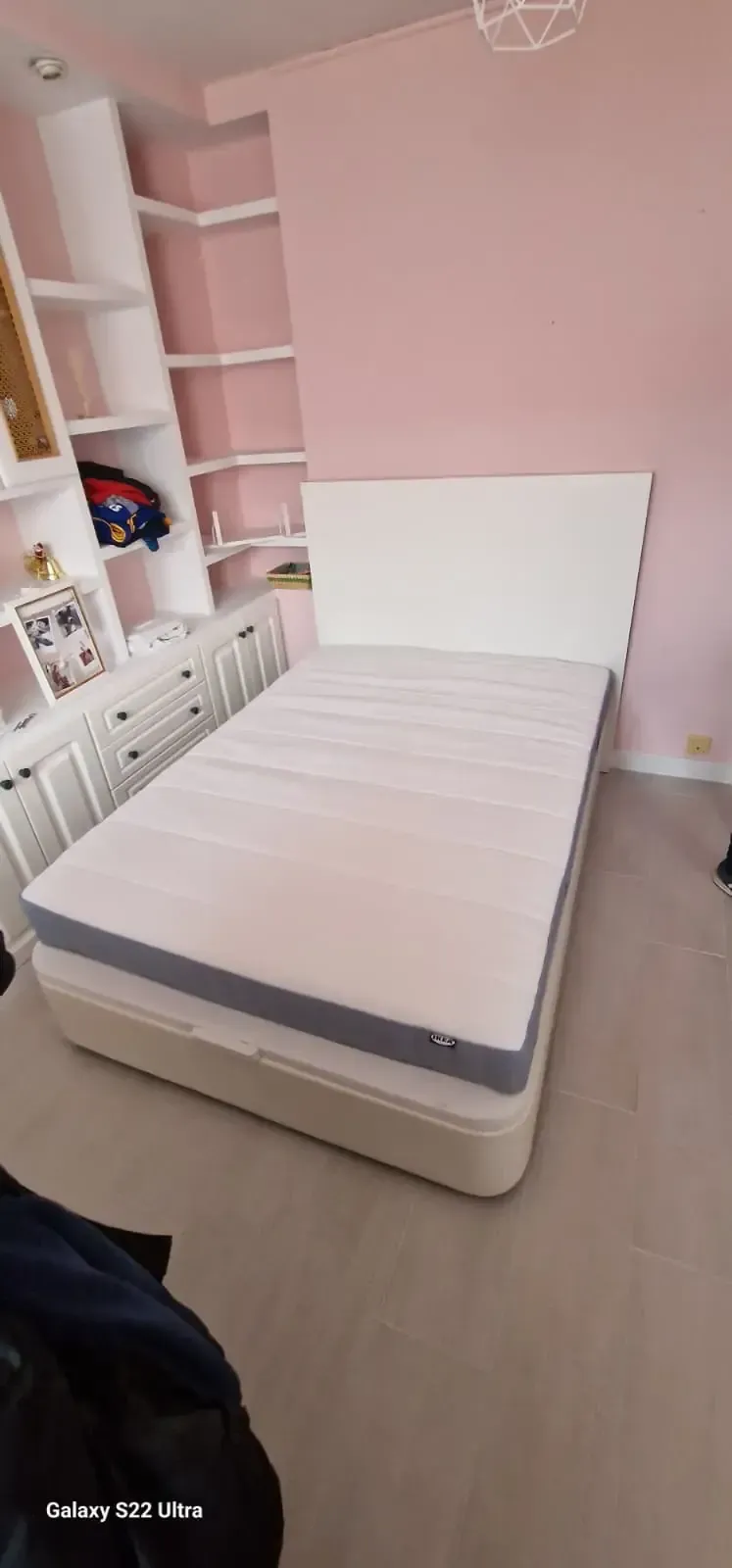 Una cama en una habitación rosa. Un cabecero y un colchón blancos descansan sobre una base beige.