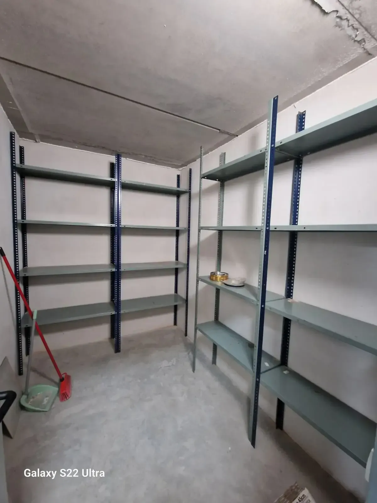 Estanterías metálicas en un almacén con paredes y techo de hormigón. Una escoba se apoya en la estantería izquierda.