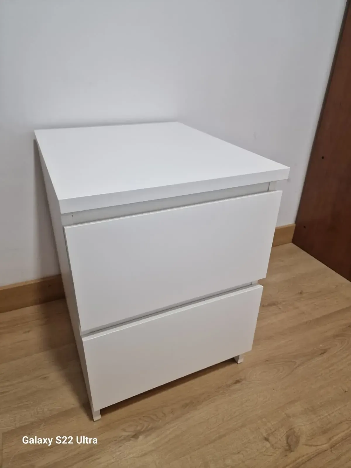 Mesita de noche blanca de dos cajones contra una pared blanca y un piso de madera.