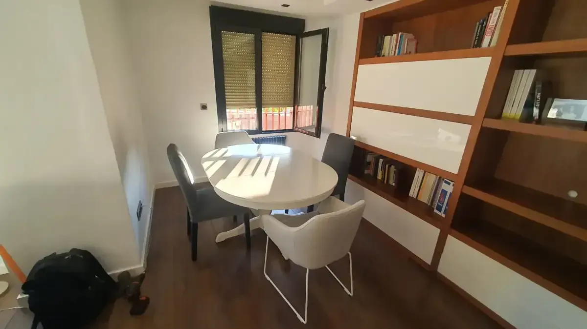 Pequeño comedor con mesa redonda blanca, tres sillas y estantería empotrada.