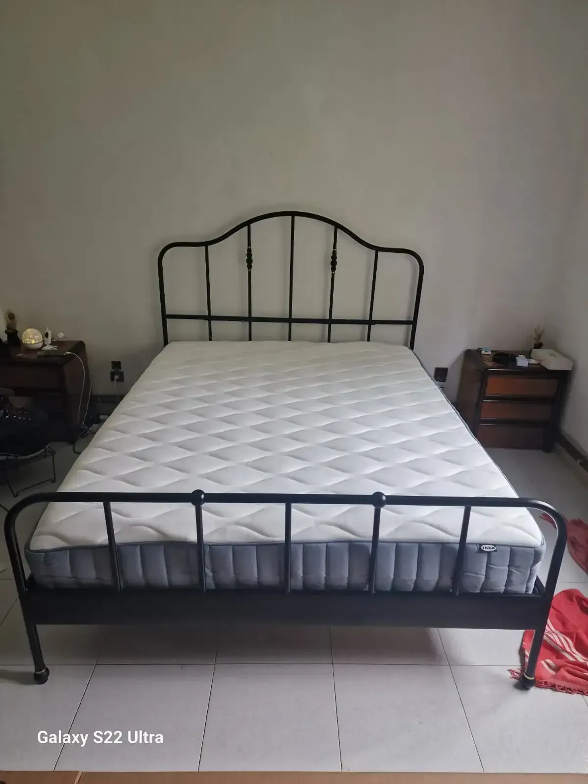 Estructura de cama de metal negro con colchón contra una pared blanca, flanqueada por mesitas de noche de madera.