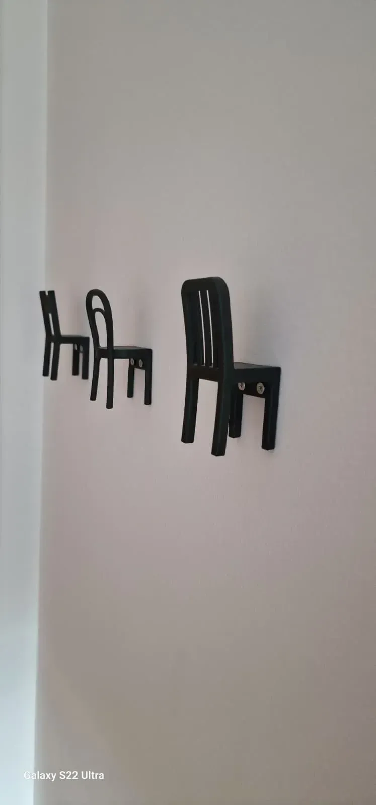 Tres ganchos de pared negros en forma de silla montados en una pared gris claro.