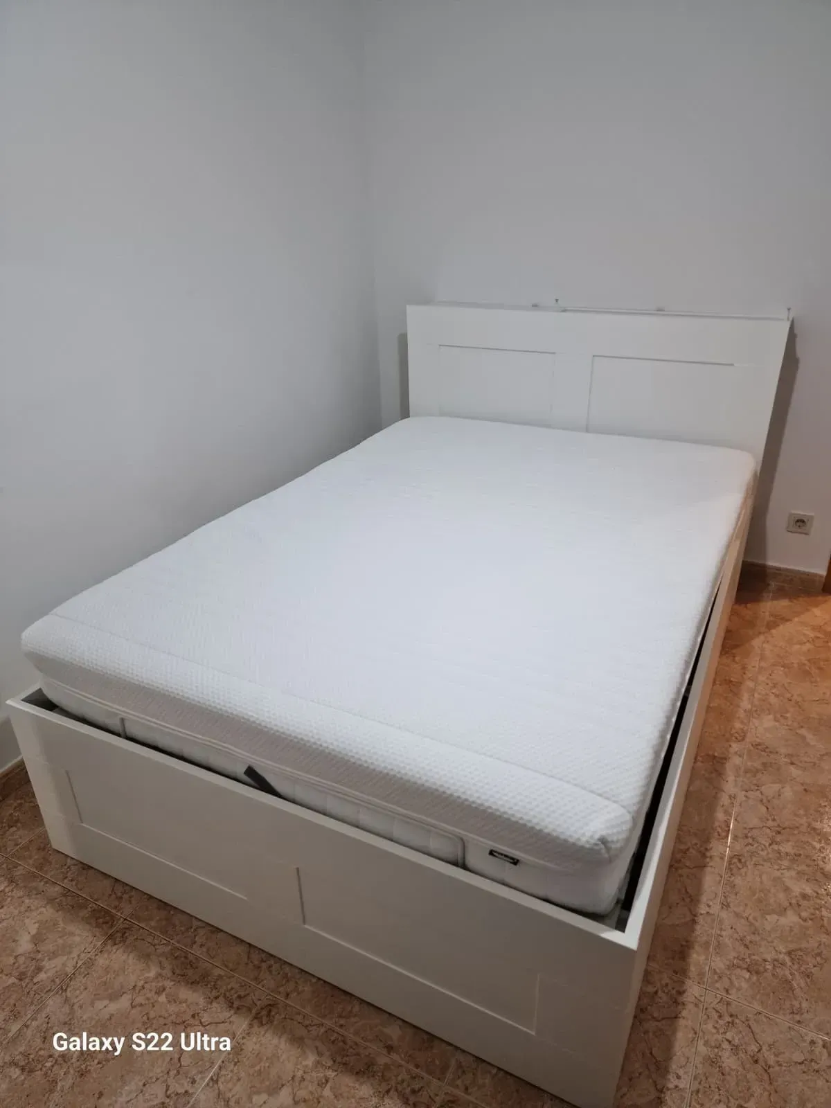 Cama blanca en esquina, con cabecero, colchón y cajones para guardar cosas.