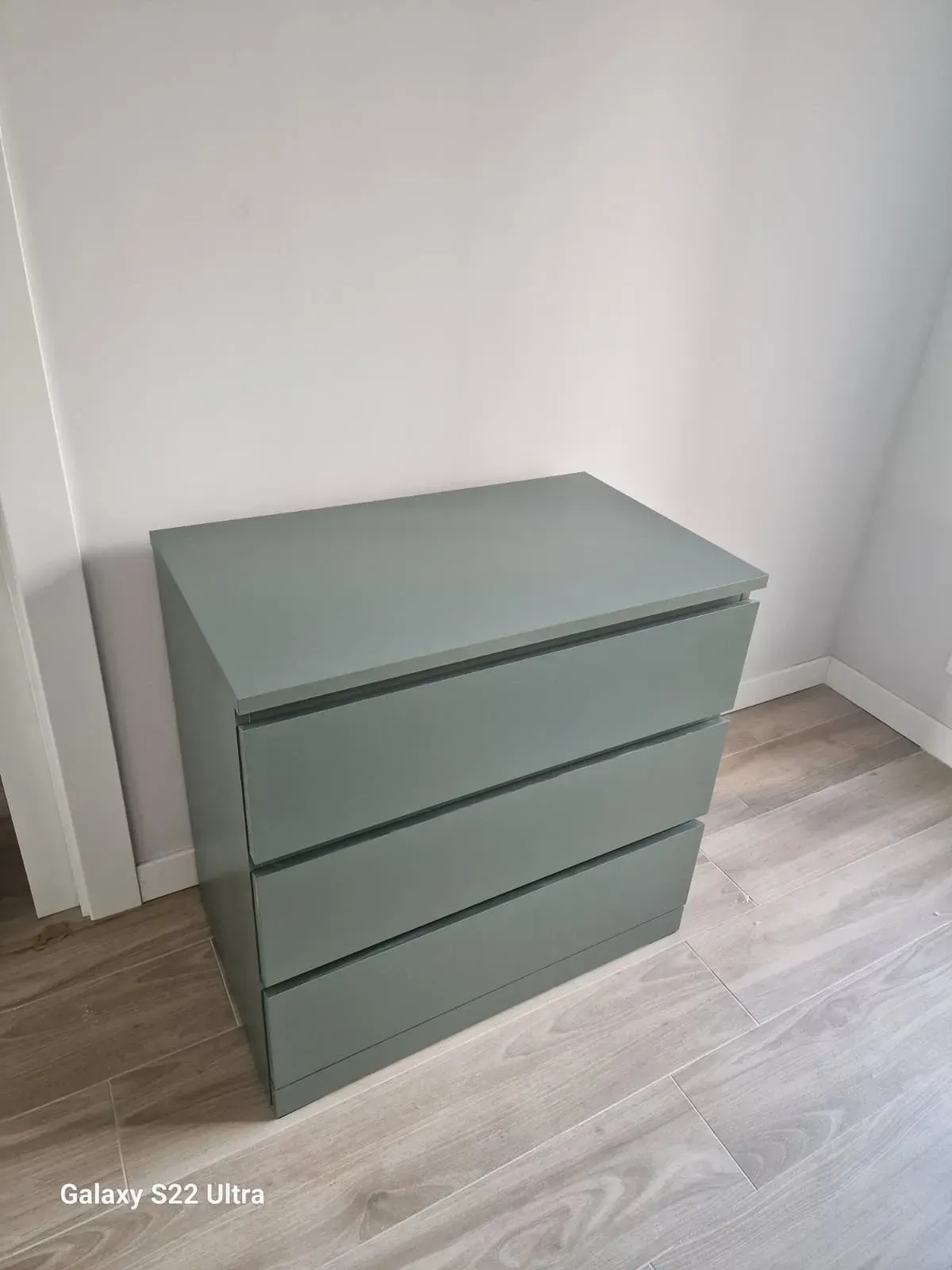 Cómoda gris de tres cajones contra una pared gris sobre un piso de madera clara.