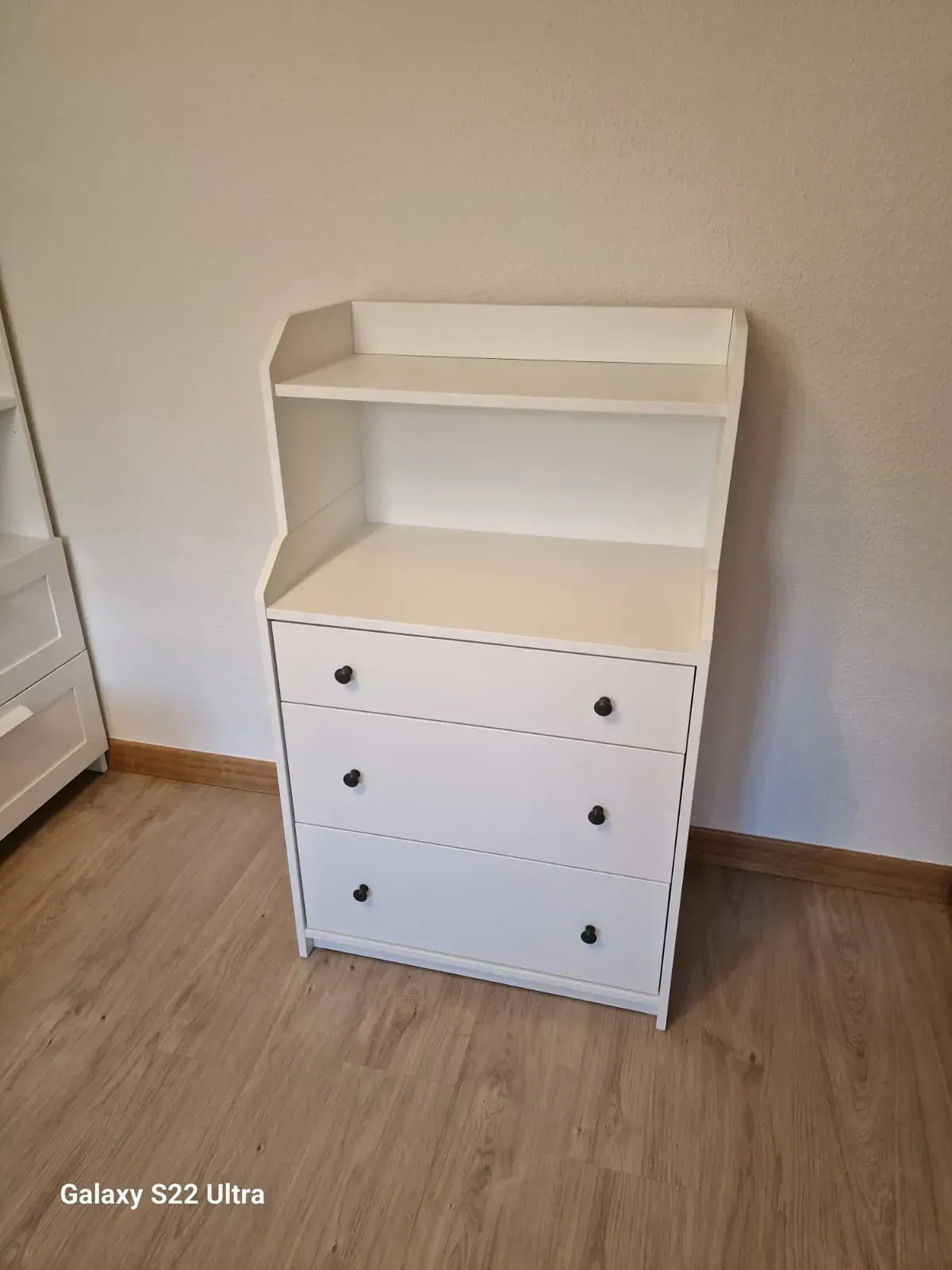 Cómoda blanca con tres cajones, un estante y tiradores negros, sobre suelo de madera.