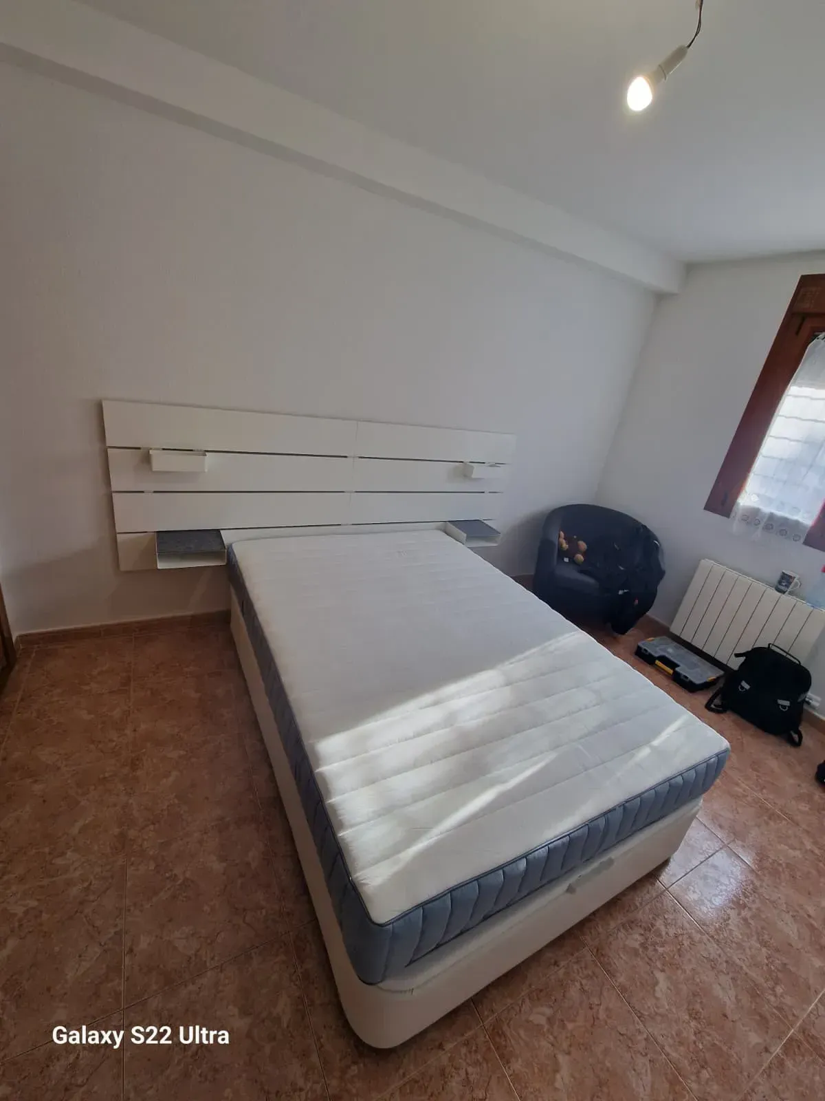 Dormitorio con cama blanca, cabecero con pequeños estantes y suelo de baldosas.