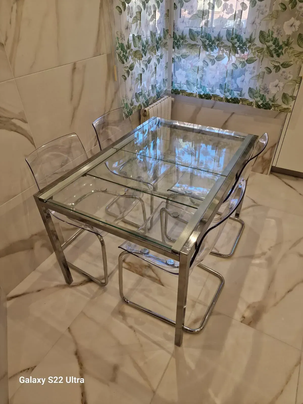 Mesa de comedor de vidrio y sillas transparentes en una habitación con pisos de mármol y una pared con estampado floral.