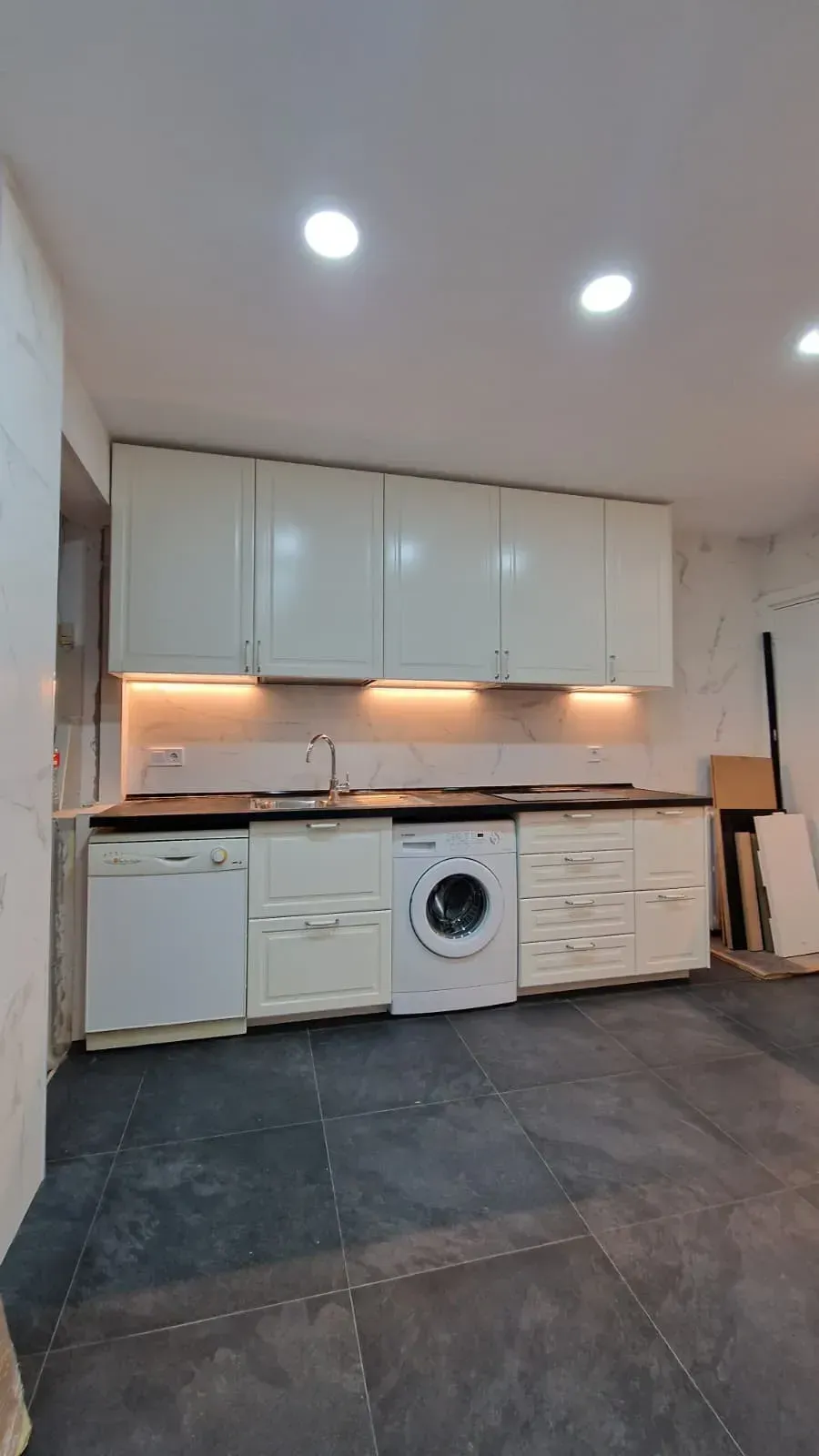 Cocina con gabinetes blancos, electrodomésticos, encimeras oscuras y pisos grises.