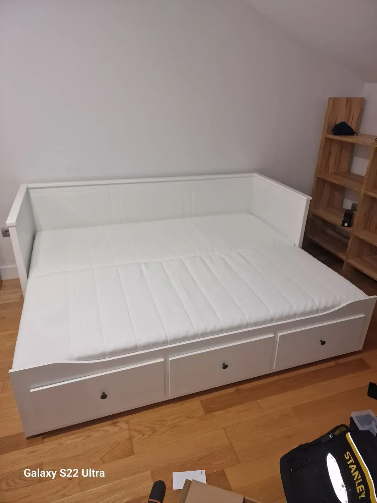 Un sofá cama blanco con tres cajones, contra una pared blanca y suelo de madera. A la derecha hay un estante de madera.