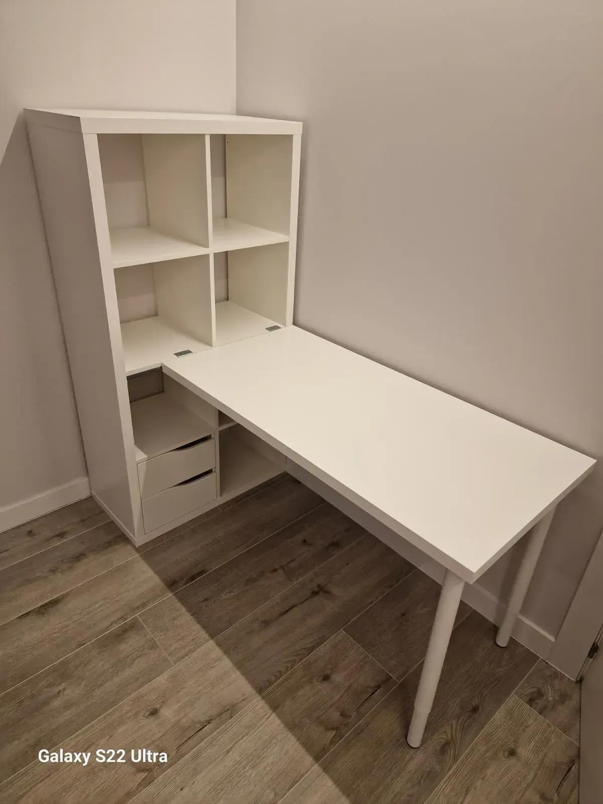 Escritorio blanco con estantes adjuntos en una esquina. Suelo de madera.