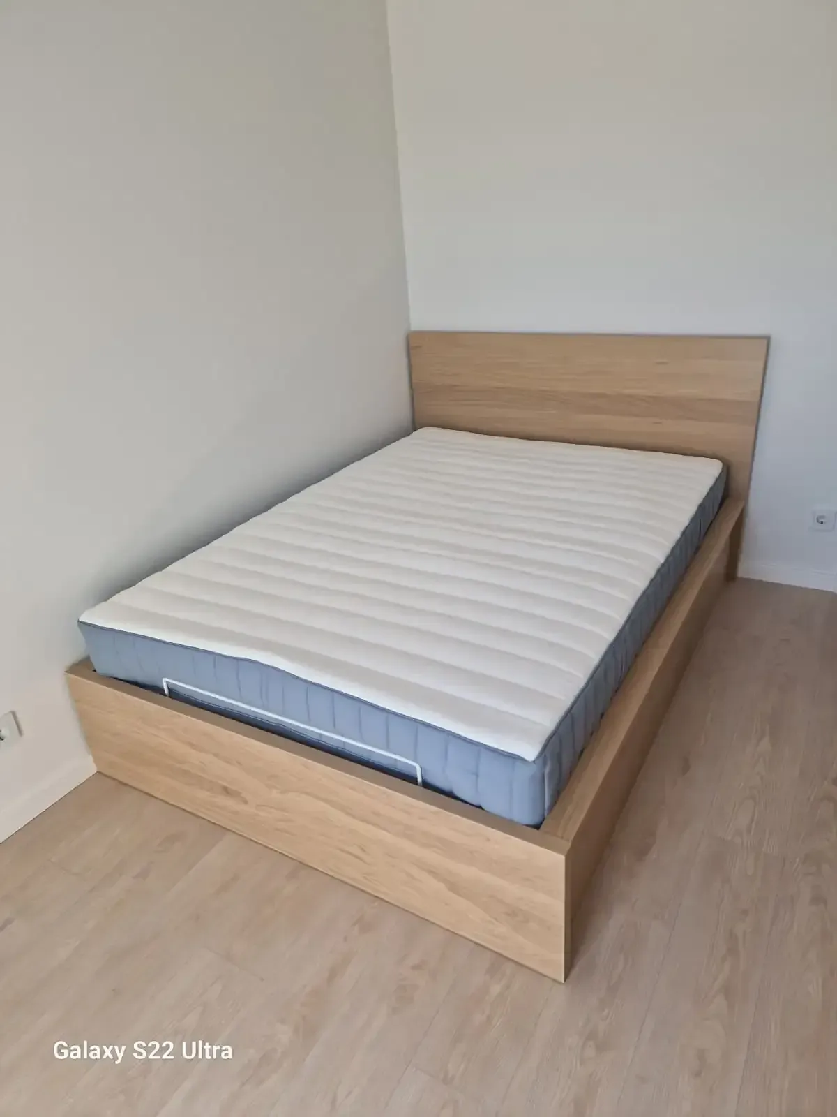 Un marco de cama de madera de color claro con un colchón en una habitación con pisos de madera clara y paredes blancas.