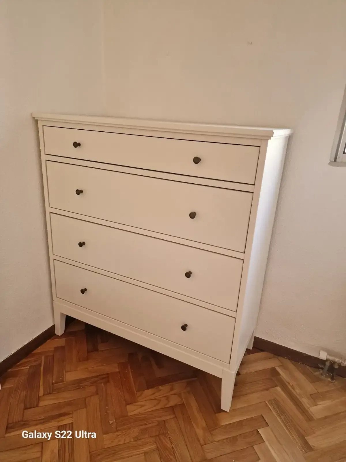 Cómoda blanca con tiradores negros, situada contra una pared blanca en una habitación con suelo de parquet.