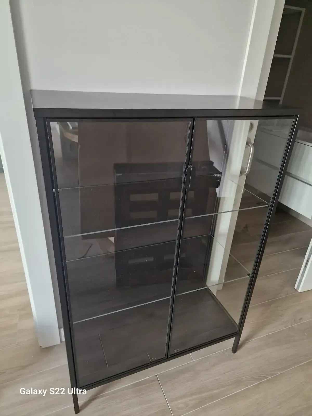 Vitrina de metal negro y cristal con puertas corredizas, colocada sobre una pared y un suelo de color claro.