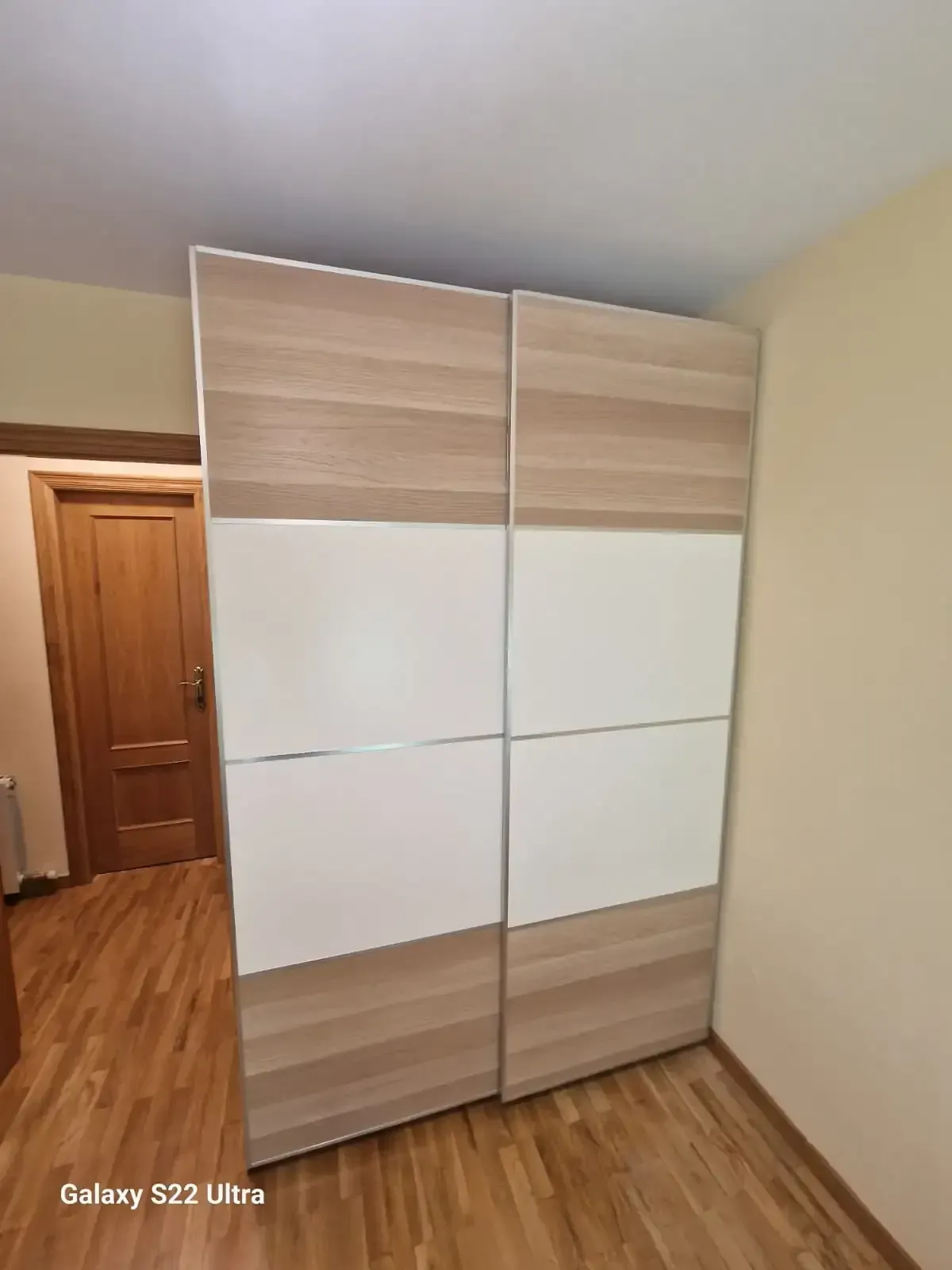 Armario corredizo de dos puertas con paneles de madera clara y blanco en una habitación con suelo y puerta de madera.