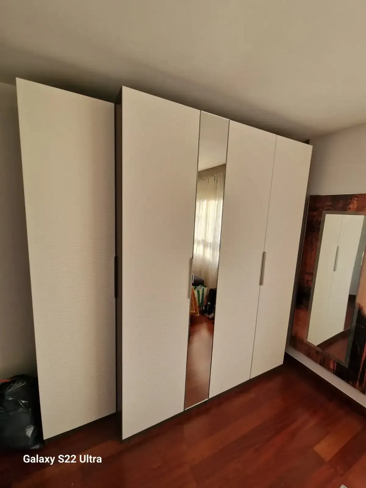 Armario blanco con puertas de espejo en una habitación con suelo de madera y un gran espejo en la pared.