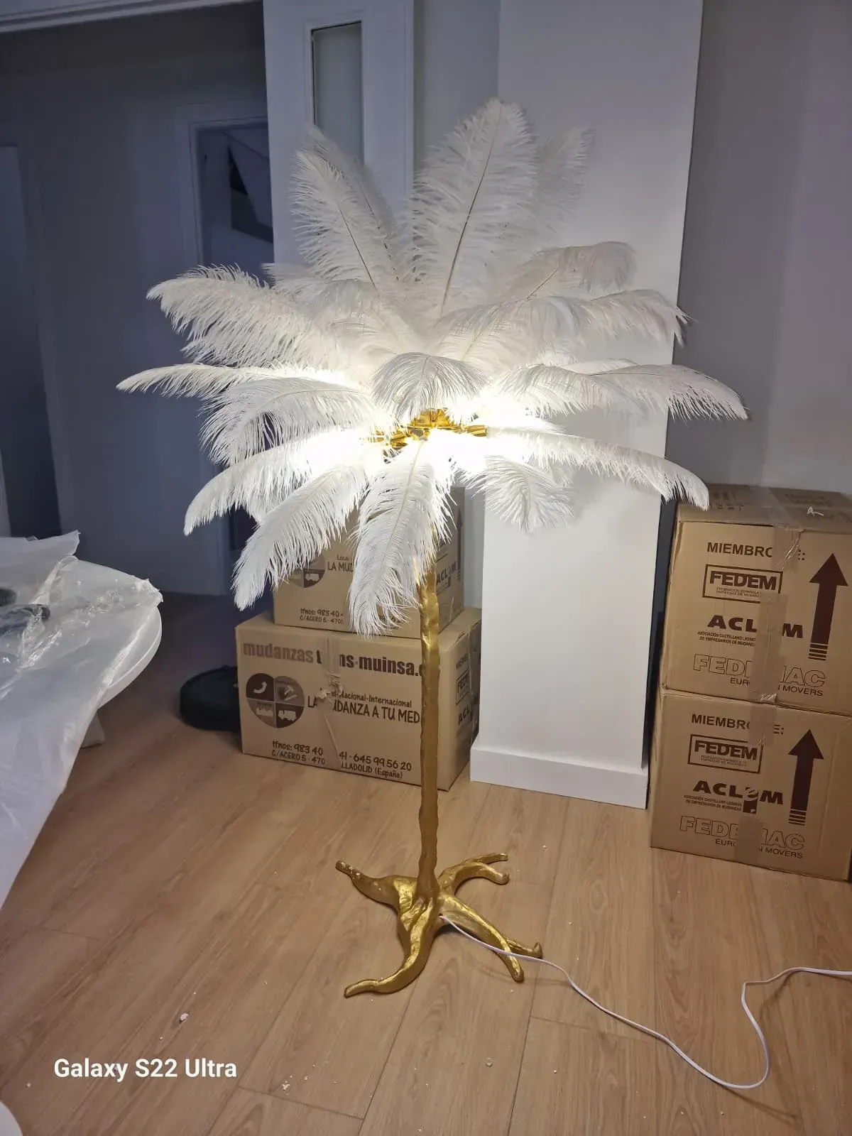 Lámpara de pie de plumas blancas con base y pie dorados, para interior sobre suelo de madera.