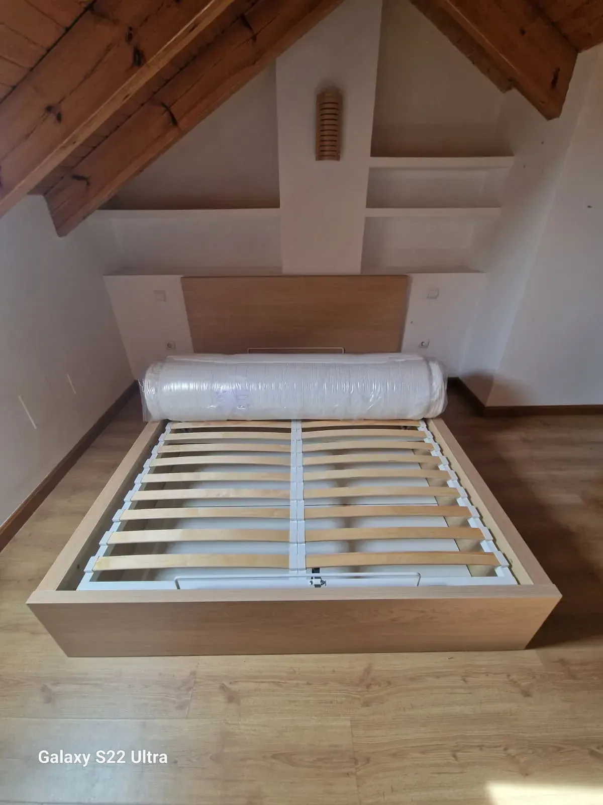 Estructura de cama con colchón en habitación abuhardillada, vigas de madera y estanterías.