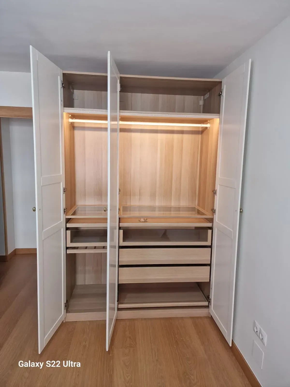 Armario blanco con puertas abiertas, estantes interiores de madera, cajones y barra con iluminación cenital.