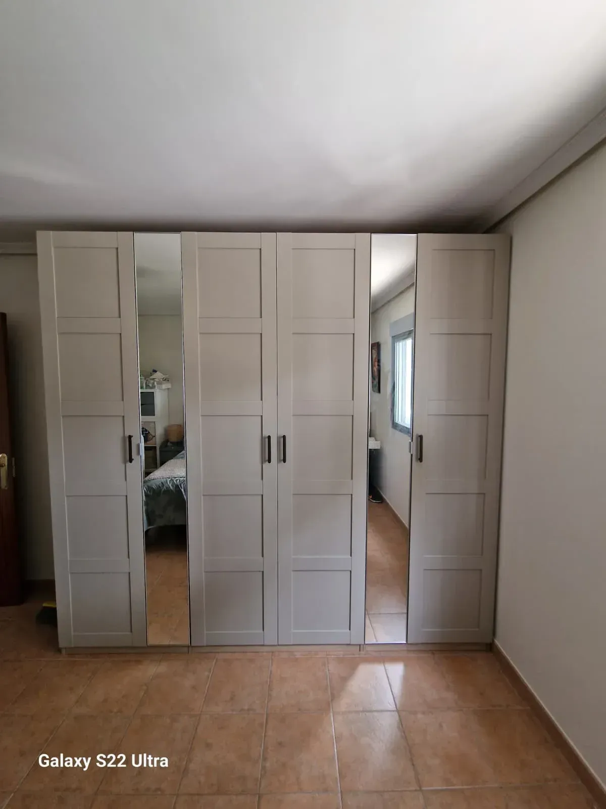 Amplio armario de color gris claro con puertas de espejo, en una habitación con suelo de baldosas y ventana.