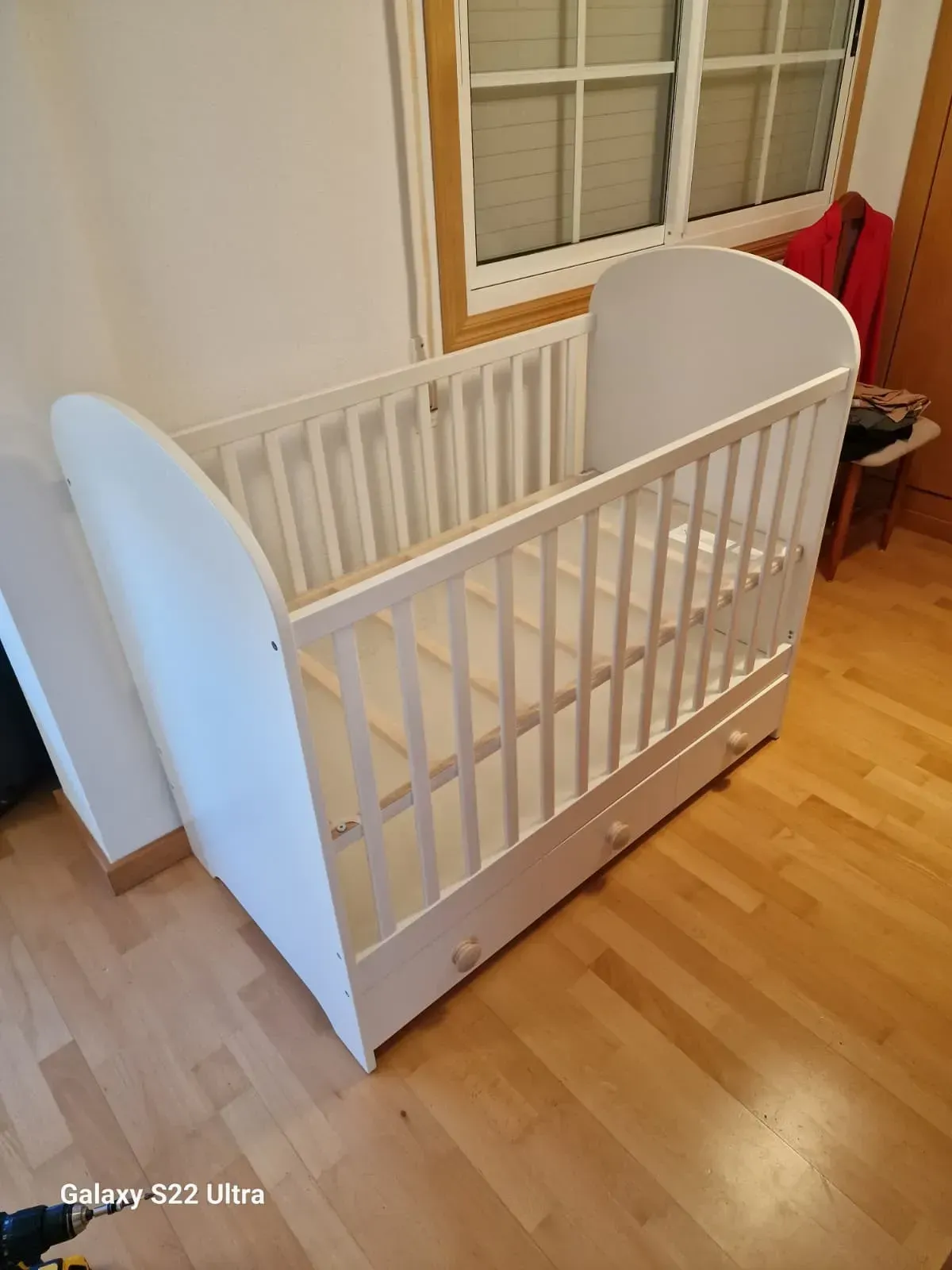 Cuna de bebé blanca con dos cajones, situada contra una pared con ventana.