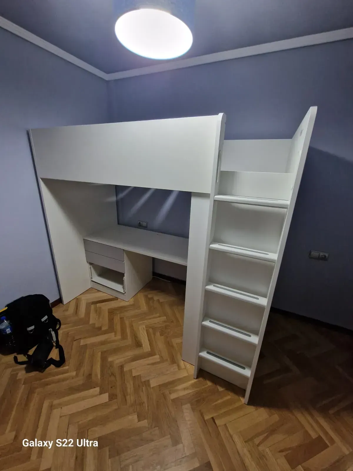 Cama alta blanca con escritorio y escalera en una habitación con suelo de madera y paredes azules.