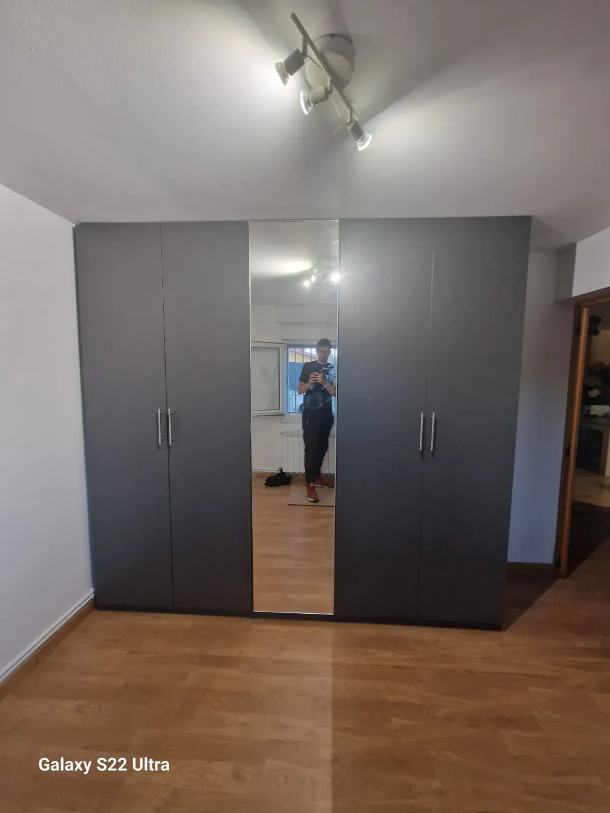 Armario gris con panel central con espejo; persona reflejada en el espejo.