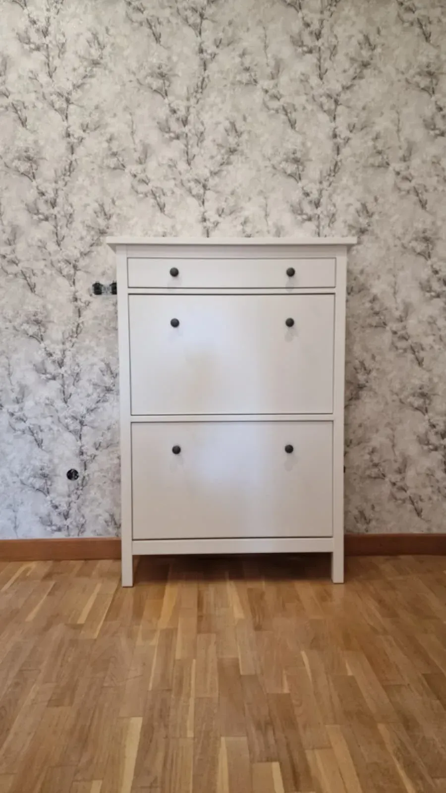 Zapatero blanco con tiradores negros contra una pared estampada, sobre un suelo de madera.