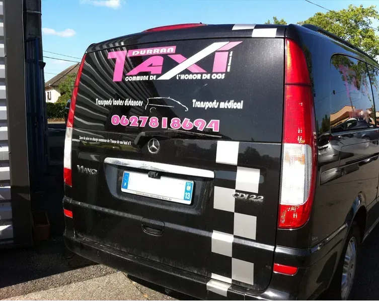 Un taxi noir de type minibus, avec l’inscription « TAXI » en rose, est garé dans une rue en plein jour.