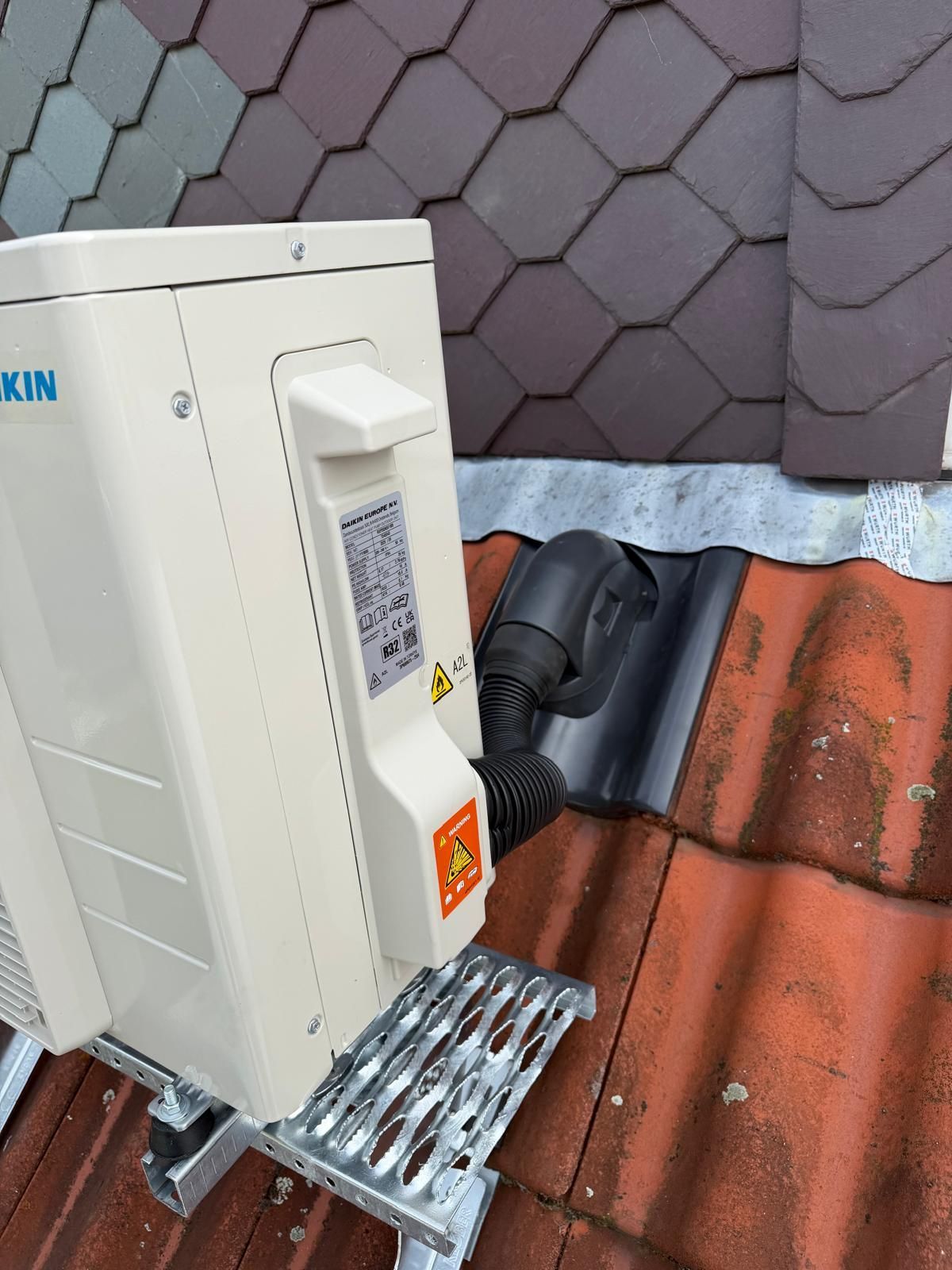 Außengerät der Klimaanlage, montiert neben einer gefliesten Wand und Dachziegeln, mit einer Metalltreppe und einem angebrachten Schlauch.