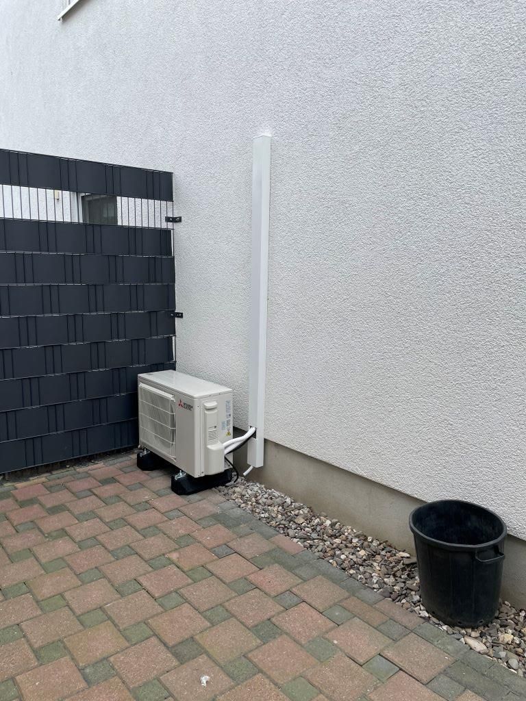 Eine weiße Klimaanlage steht auf einem gepflasterten Gehweg neben einem Zaun.