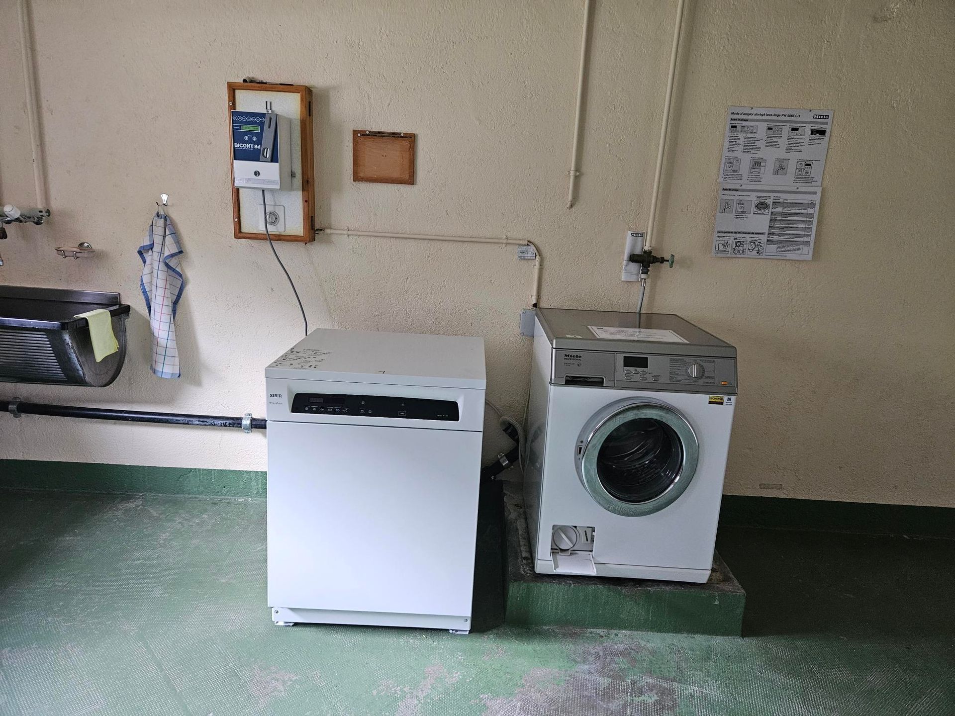 Dans une buanderie se trouvent une machine à laver et un sèche-linge, avec un compteur électrique et des instructions affichées au mur.