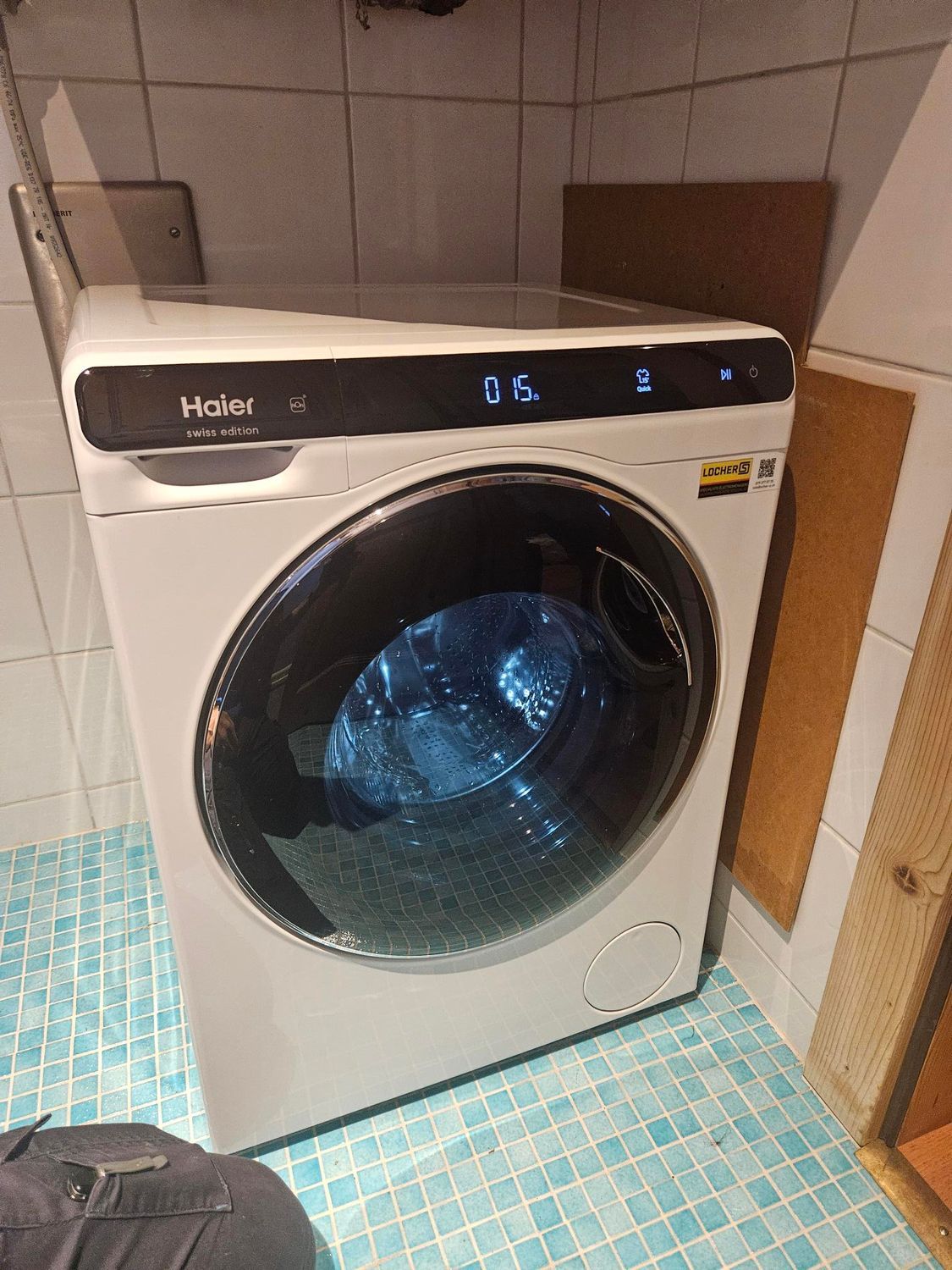 Machine à laver Haier blanche dans une salle de bains avec sol carrelé bleu.