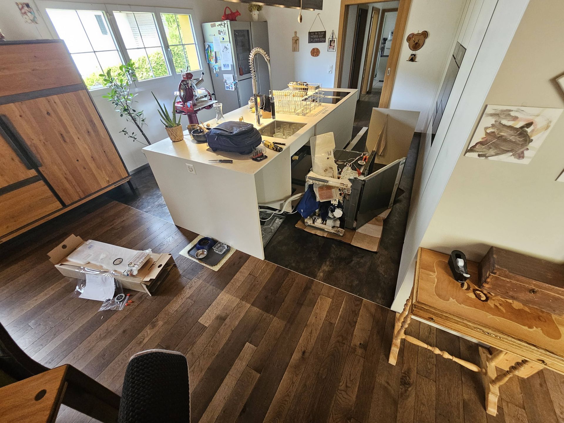 Cuisine encombrée avec un îlot blanc, divers objets éparpillés sur le sol, les placards et la table.