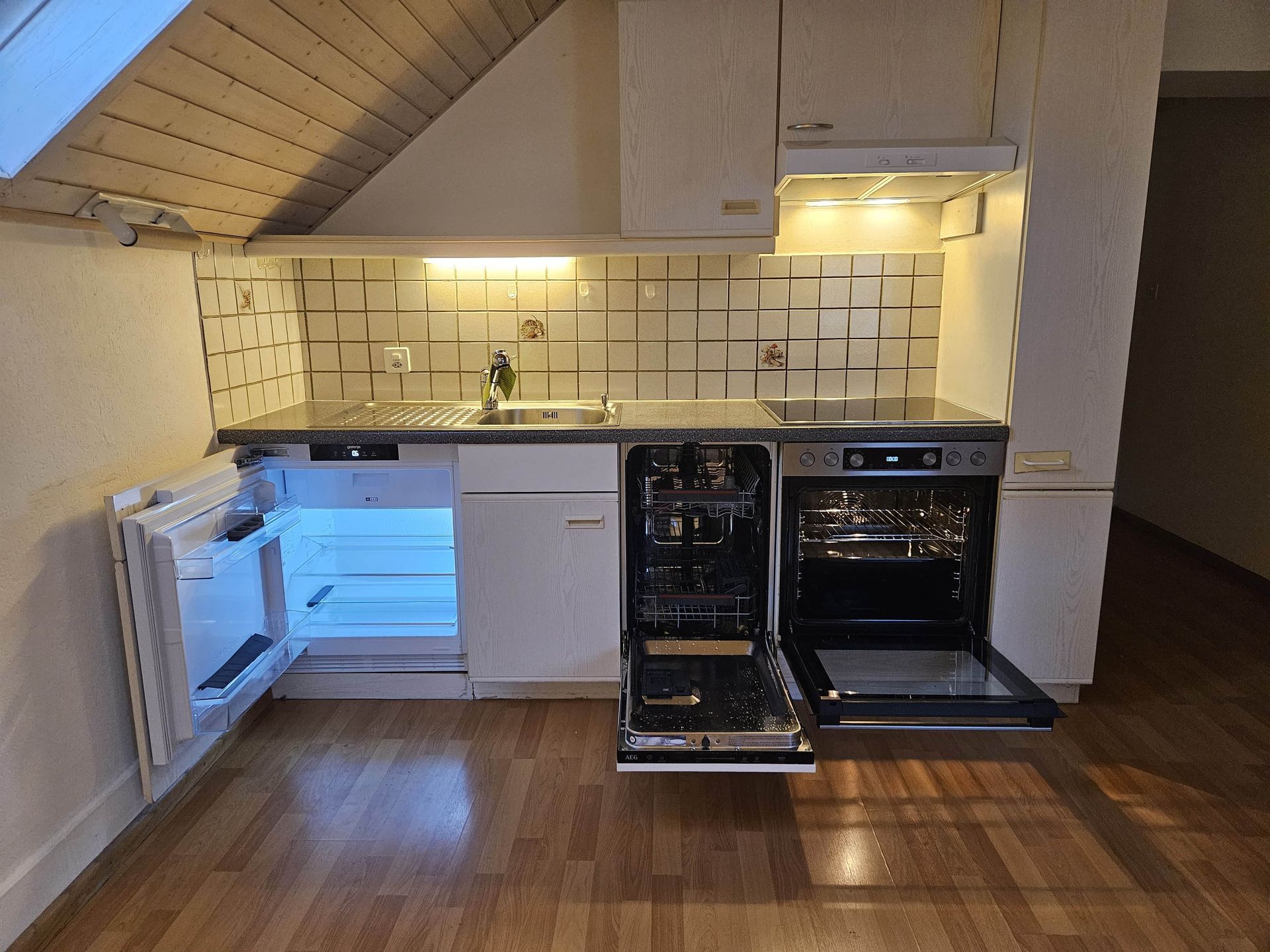 Petite cuisine avec réfrigérateur ouvert, lave-vaisselle et four. Placards beiges, crédence carrelée et parquet.