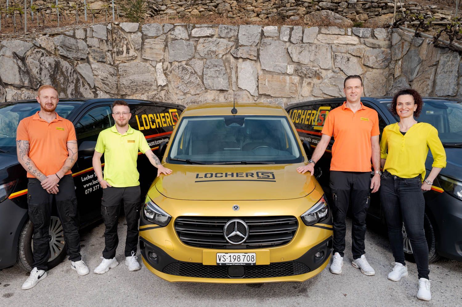 Cinq personnes se tiennent debout avec trois fourgonnettes dorées et noires, contre un mur de pierre. Des logos sont visibles.