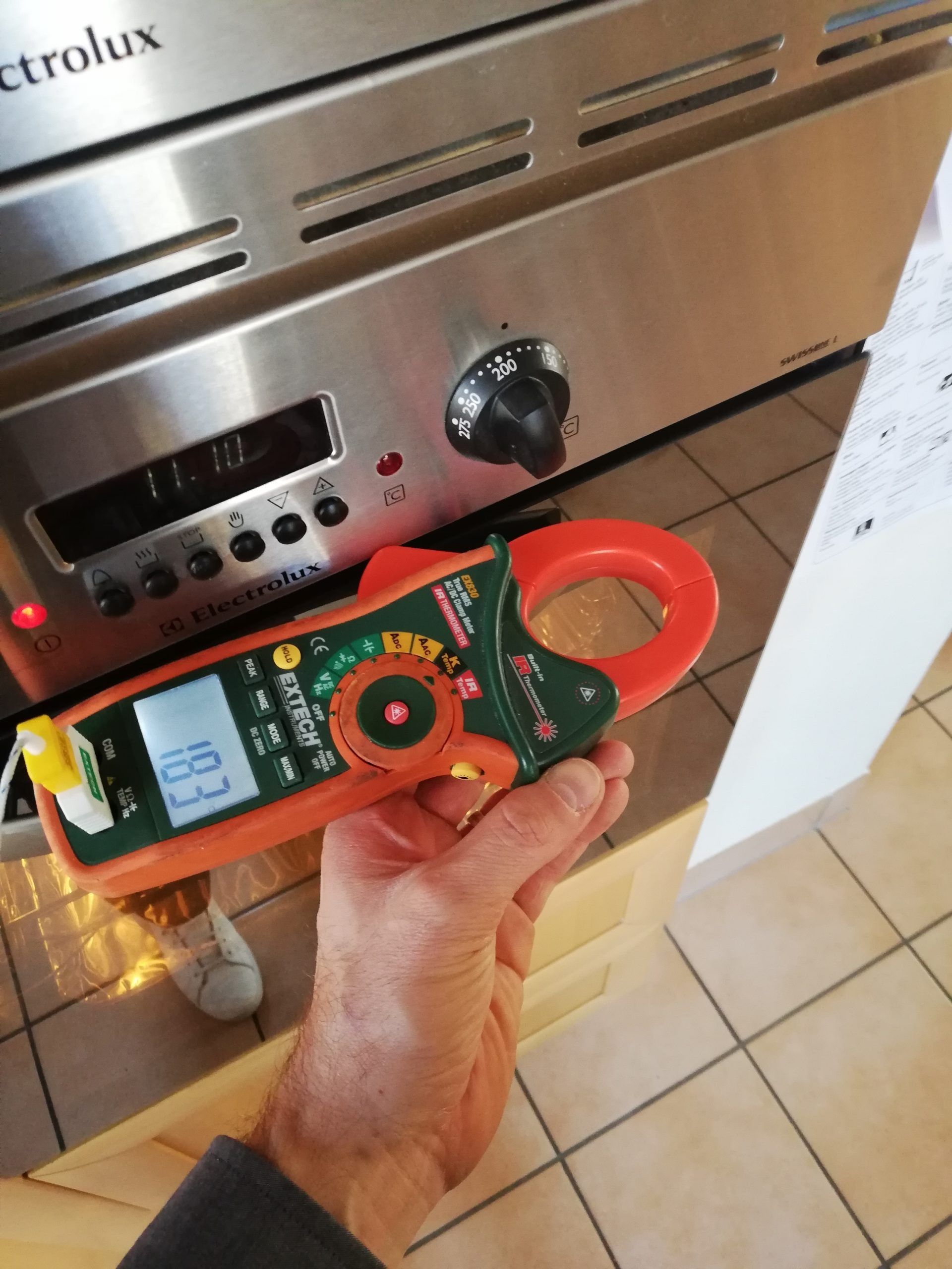 Une main tenant une pince ampèremétrique près d'un four, affichant 18,3 ampères.