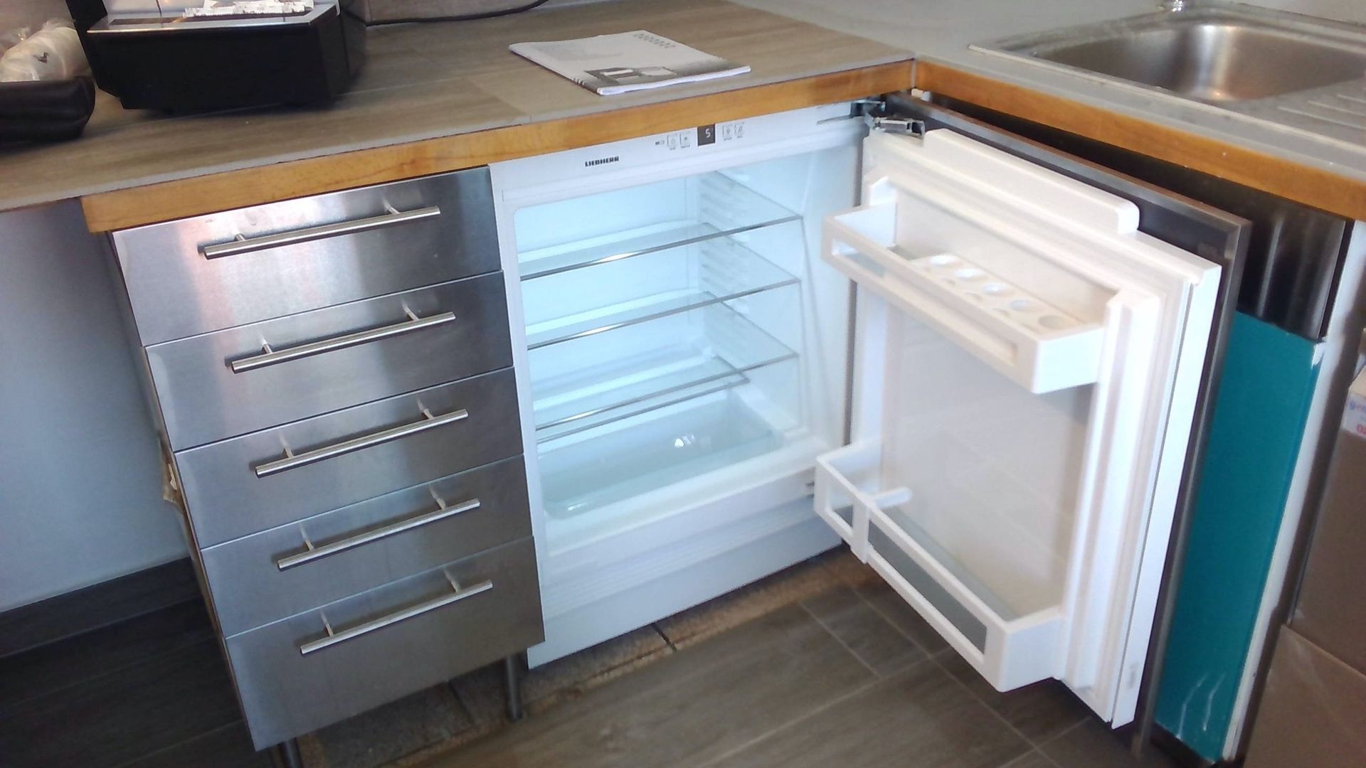 Un petit réfrigérateur à porte ouverte est intégré dans un meuble. Des tiroirs en acier inoxydable se trouvent à gauche.