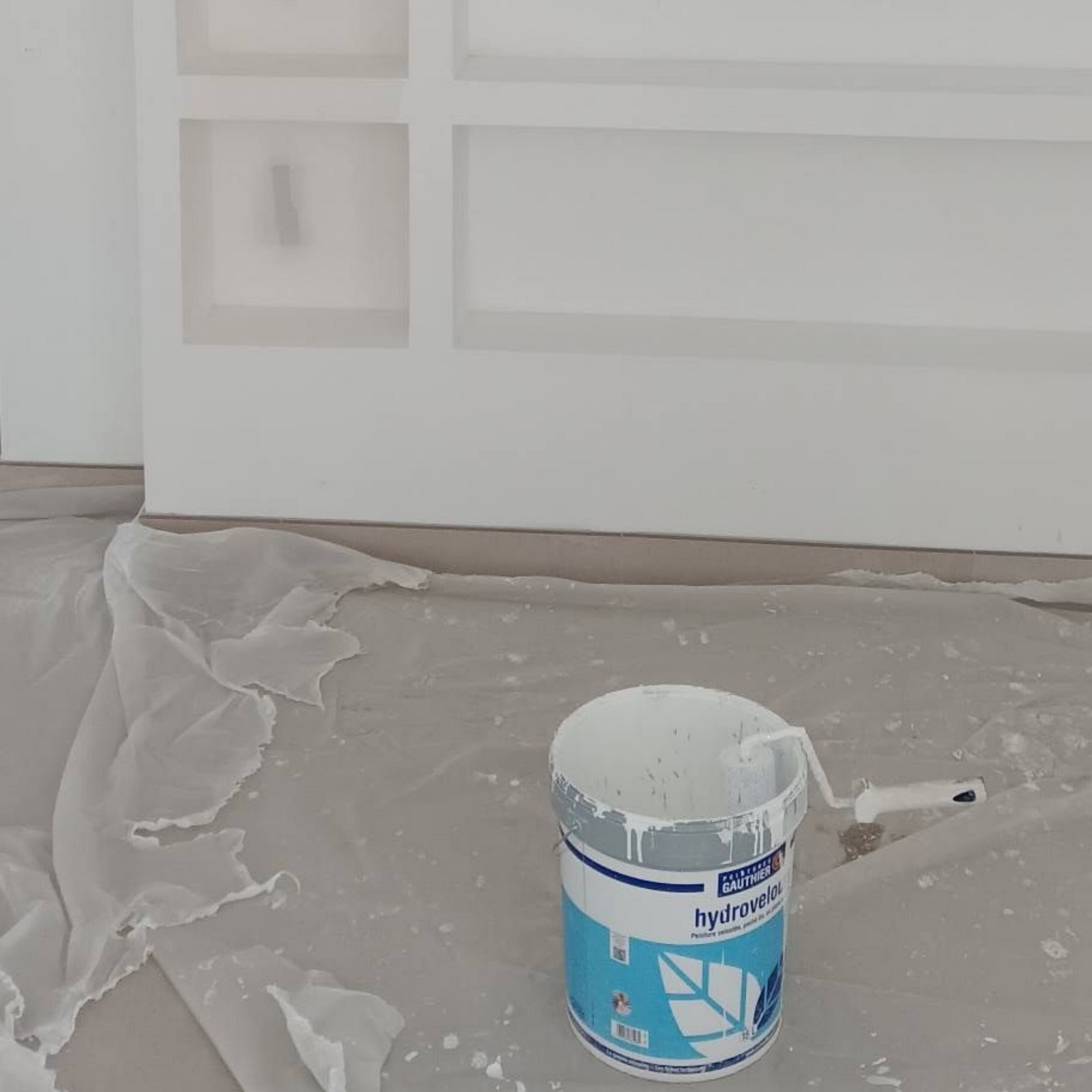 Un mur partiellement peint avec des étagères intégrées, un pot de peinture avec un rouleau posé au sol.