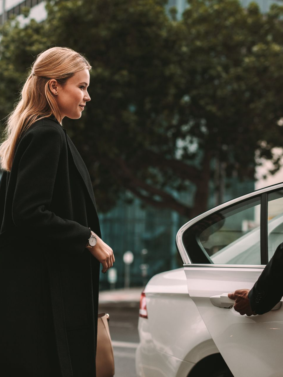 Une femme en manteau noir s'apprête à monter dans une voiture blanche, lorsqu'une personne ouvre la portière.