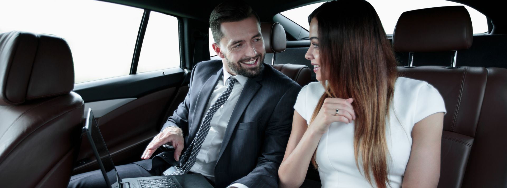 Un homme en costume regarde une femme assise dans une voiture, souriante. Ils sont tous deux assis à l'arrière. Intérieur marron.