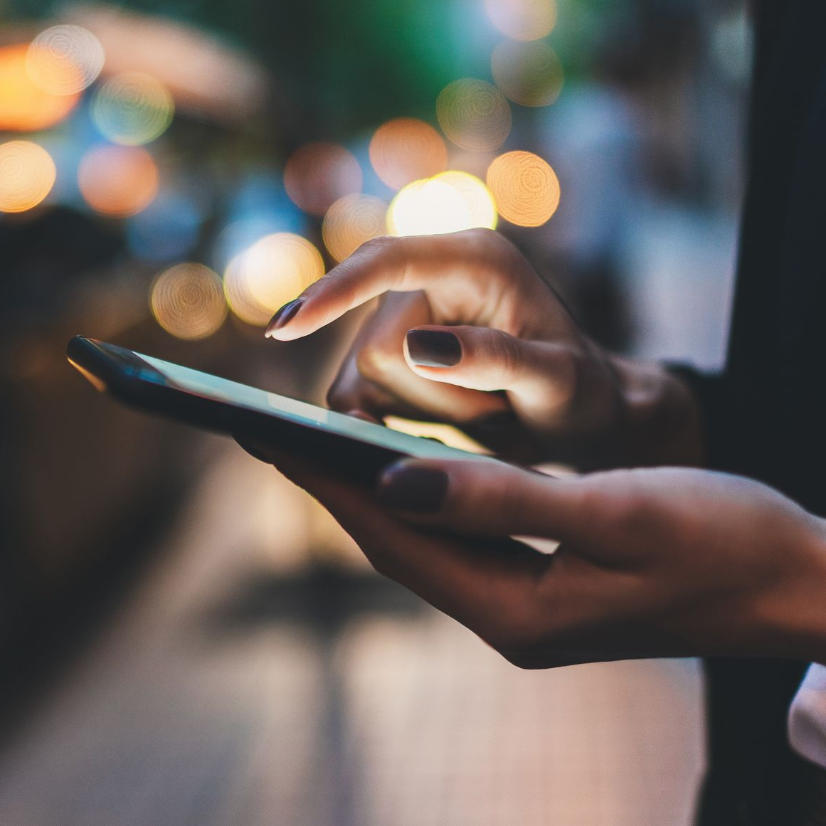 Mains d'une personne utilisant un smartphone à l'extérieur, la nuit. Du bout des doigts, l'écran se touche ; lumières floues en arrière-plan.