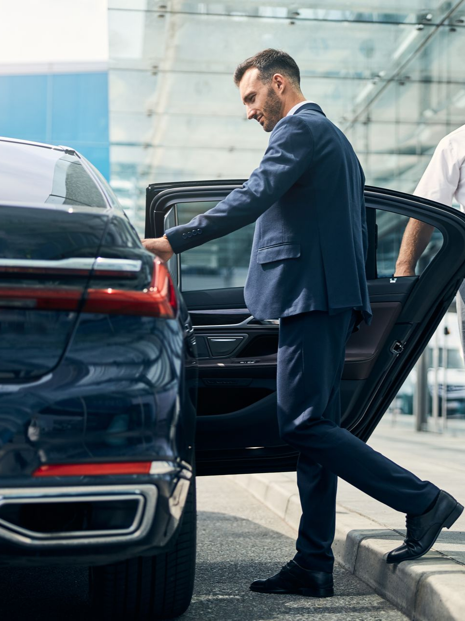 Un homme en costume monte d'une berline bleu foncé ; un portier l'assiste près d'un immeuble moderne.