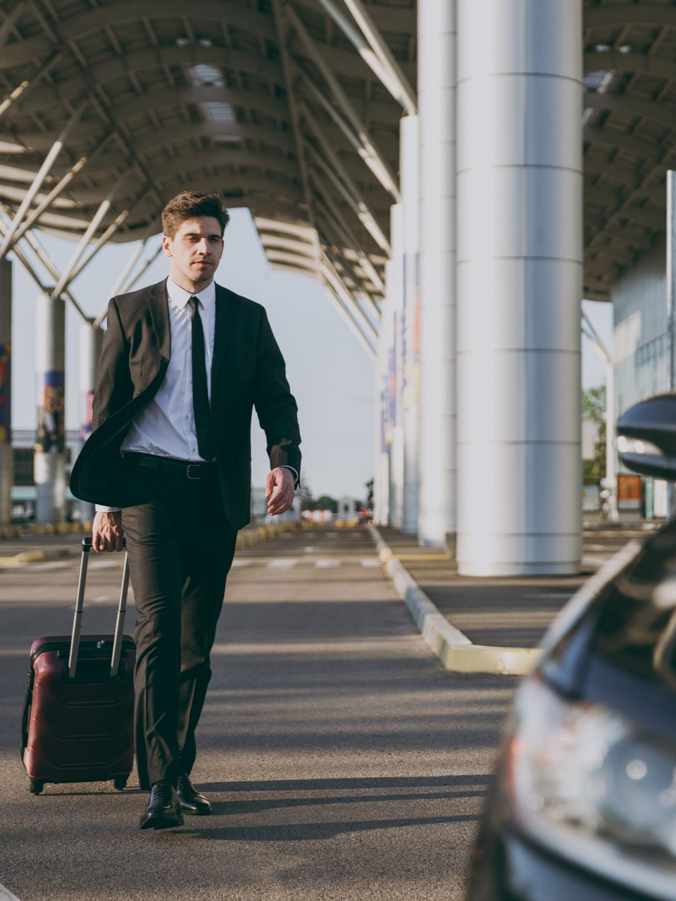 Un homme en costume marchait avec une valise à roulettes près d'une voiture, devant un immeuble.