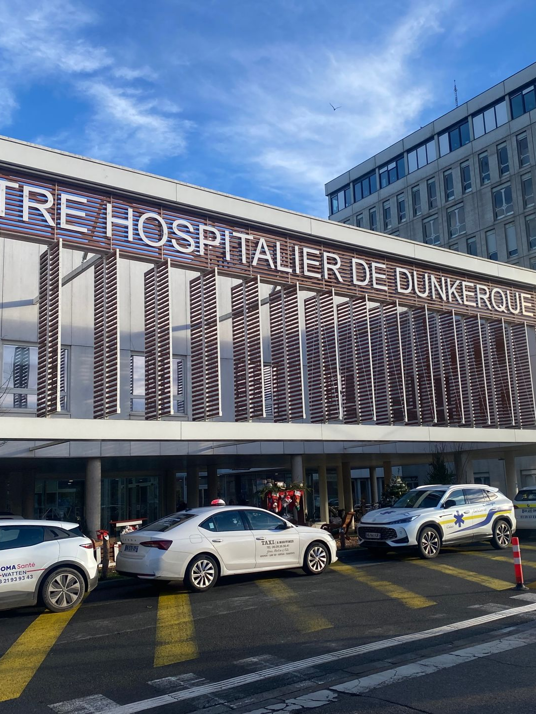 Entrée du Centre Hospitalier de Dunkerque avec des voitures garées devant, sous un ciel bleu.