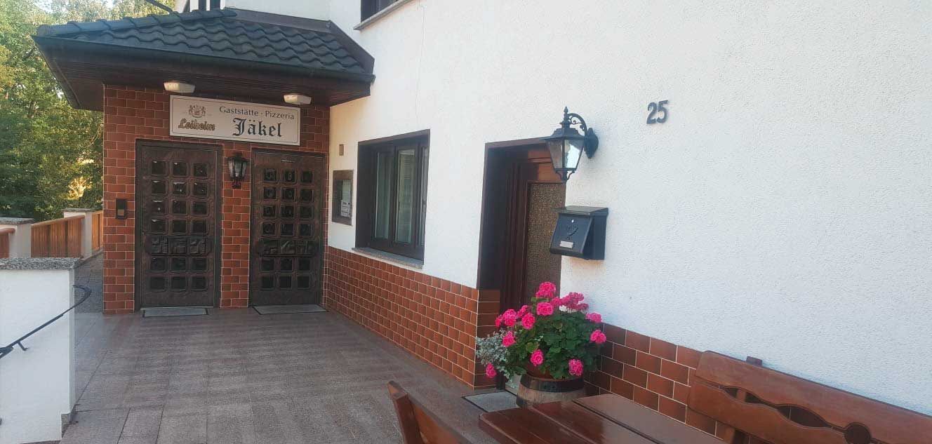 Die Außenfassade des Restaurants besteht aus Ziegelsteinen und weißen Wänden, Holzverzierungen und einem Blumentopf. An der Wand steht die Hausnummer 25.