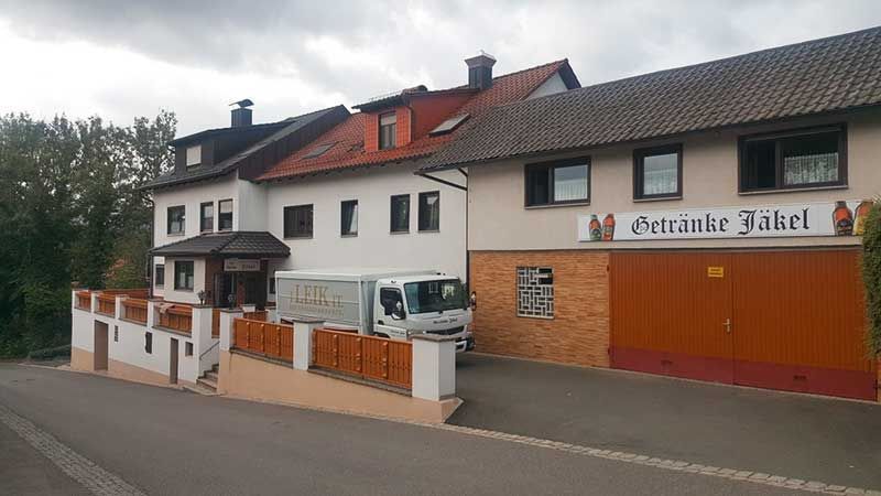 Gebäude mit einem davor geparkten LKW; Schild mit der Aufschrift „Benzinlie Jakel“. Braune Garagentore, grauer Himmel.