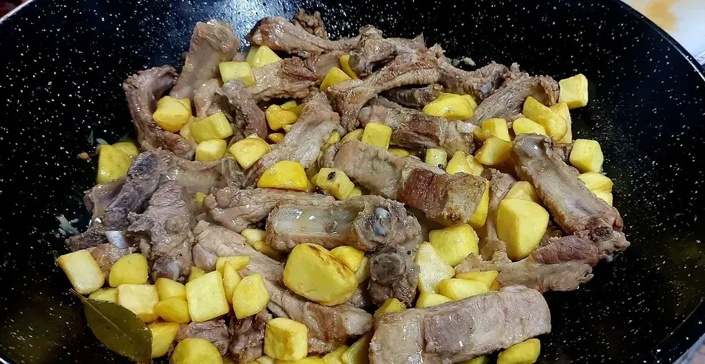 Una sartén oscura moteada contiene costillas de cerdo cocidas mezcladas con patatas fritas cortadas en dados y de color dorado, y una hoja de laurel.