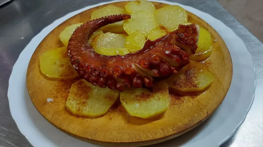 Un tentáculo de pulpo cocido servido sobre una cama de patatas en rodajas y sazonadas en un plato de madera.