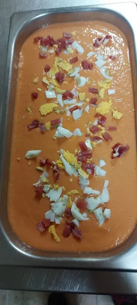Una bandeja rectangular de metal llena de salmorejo, una sopa fría de tomate y naranja, cubierta con huevo duro picado y jamón.