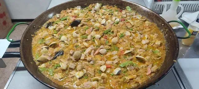 Una gran paellera redonda de metal llena de paella de marisco recién hecha, con almejas, trozos de pescado y verduras verdes.